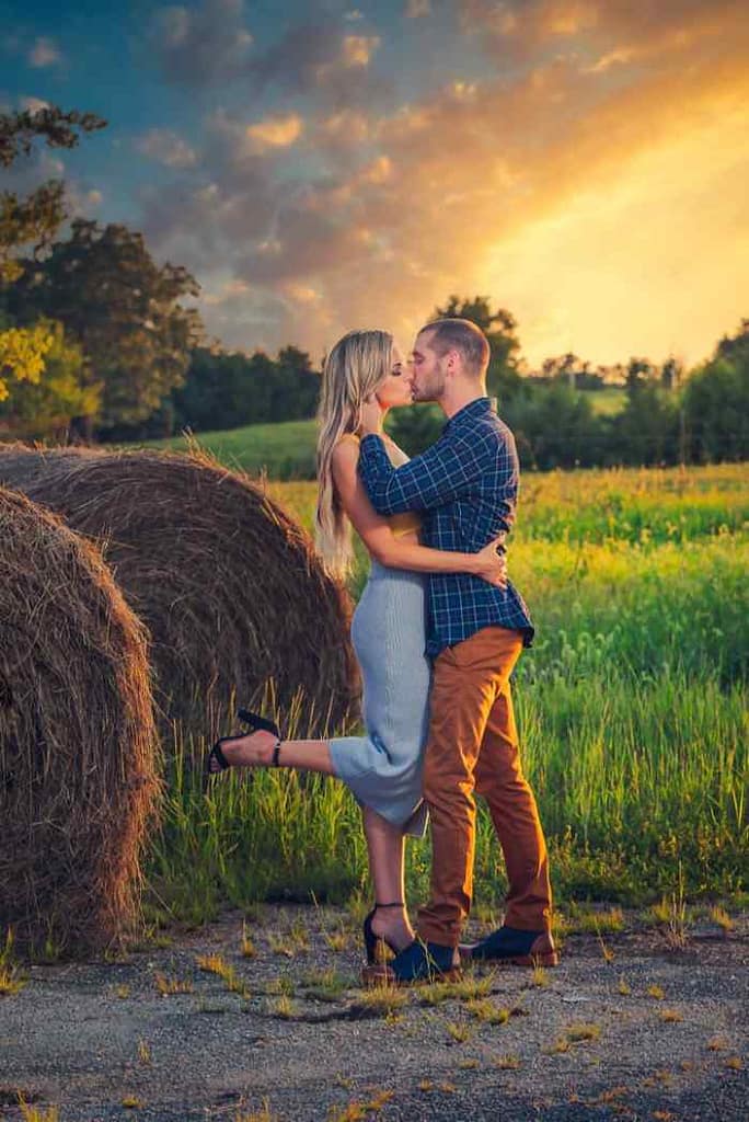 Couple in an engagement photo shoot in a hay field by some big bales of hay